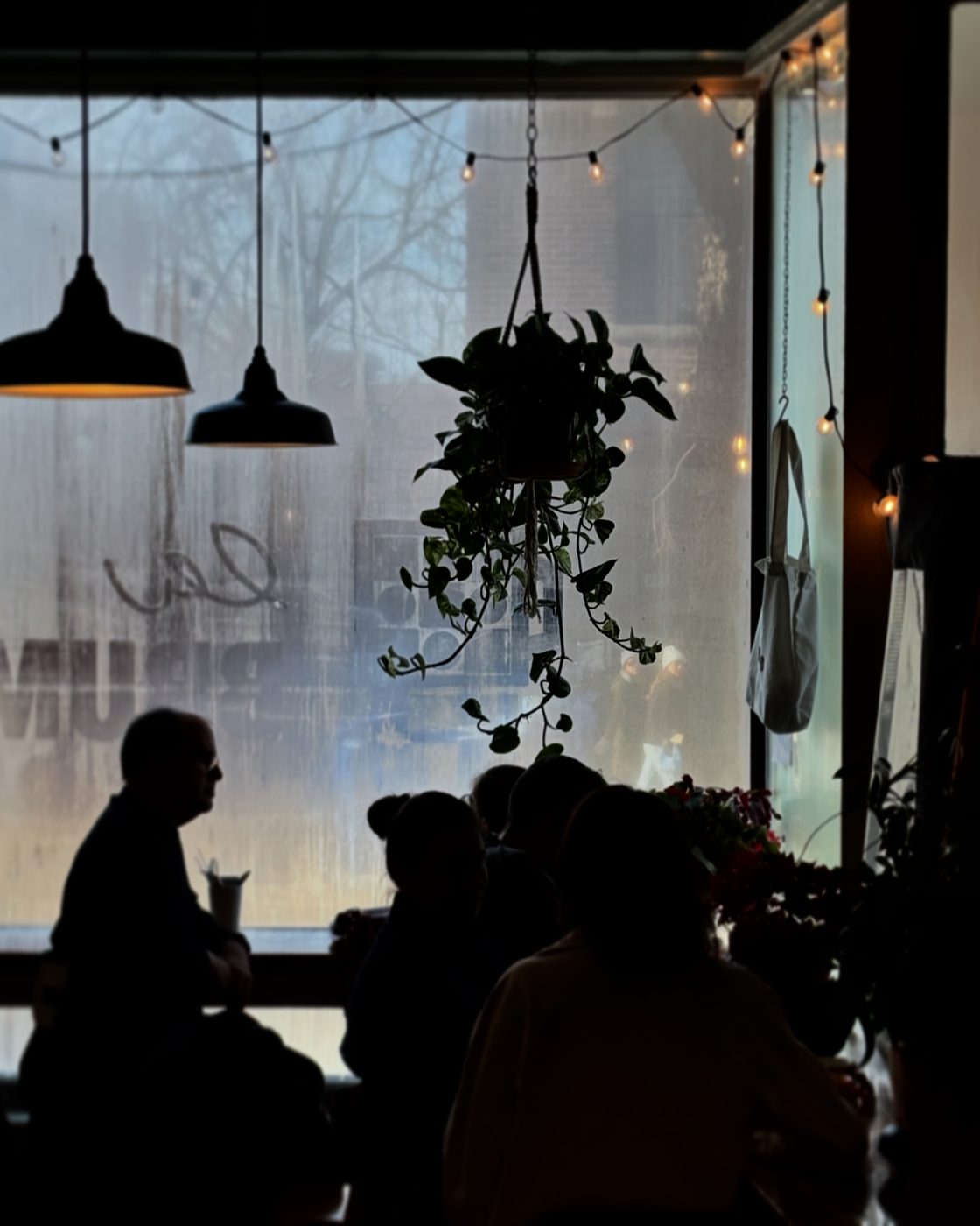 Silhouettes de clients devant la grande vitrine embuée de brume.café, sous une plante suspendue et des guirlandes lumineuses